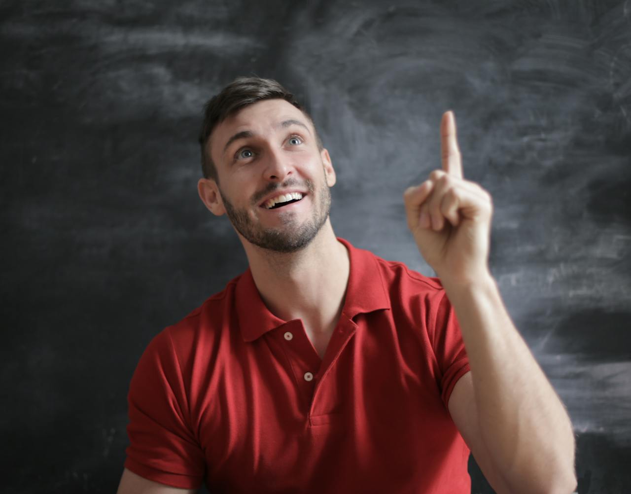 Services Happy young man in a red polo shirt smiling and pointing upwards in front of a blackboard.